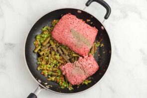 A skillet with cooked vegetables and raw ground beef on top.