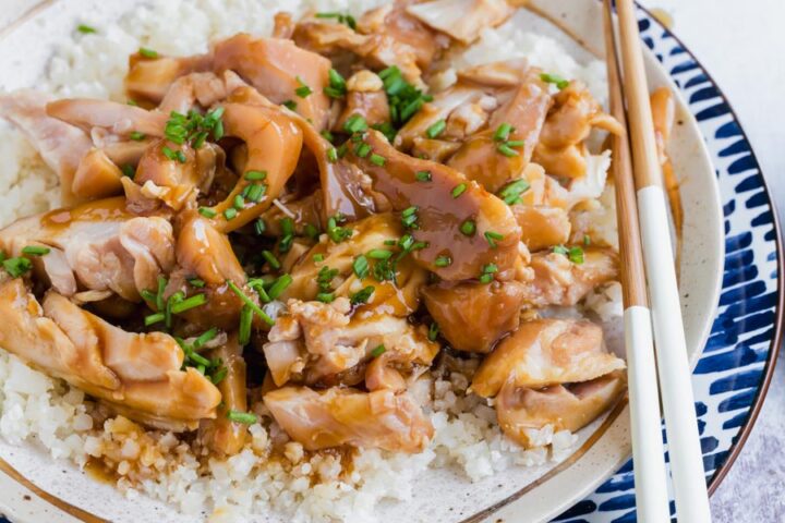 Close-up of tender chicken pieces in savory sauce over cauliflower rice with chopsticks alongside.