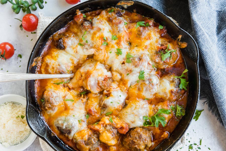 looking down on a cast iron skillet filled with meatballs in a pasta sauce covered with cheese with tomatoes and parmesan cheese near