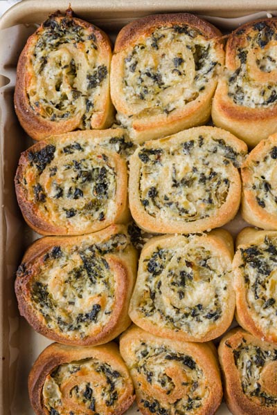 Rows of spinach and cheesy pinwheel buns baked to golden brown and melted cheese on top on a baking pan.
