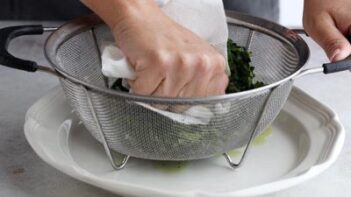 A hand pressing a paper towel into cooked, wilted spinach in a colander sitting on a plate in order to squeeze the moisture out.