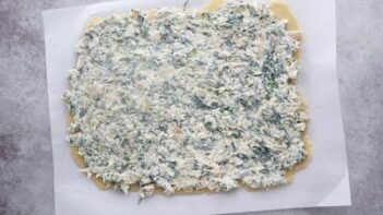Spinach artichoke spread on top of pastry dough sitting on a sheet of parchment paper.