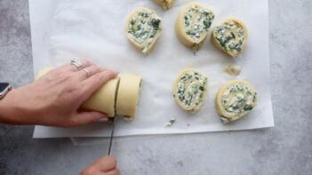 A hand slicing through a log of rolled spinach and pastry dough, cutting them into slices.