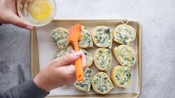 A hand brushing melted butter on top of spinach artichoke buns unbaked on a baking sheet.