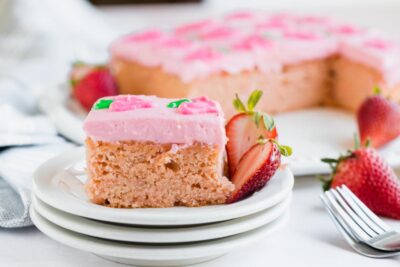 a square slice of keto strawberry cake on a stack of white plates with two sliced strawberries on the side and forks nearby