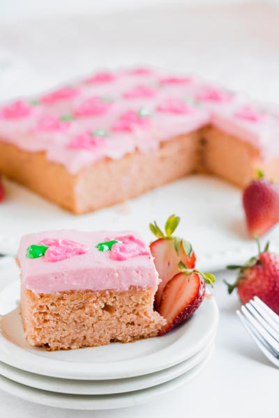 a slice of pink cake on a white plate with fresh strawberries next to it and a full sheet cake behind