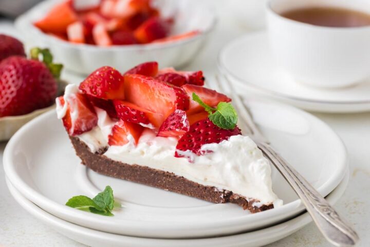 A slice of keto strawberry cream pie with a chocolate crust and fresh strawberries on a white plate beside a cup of tea.