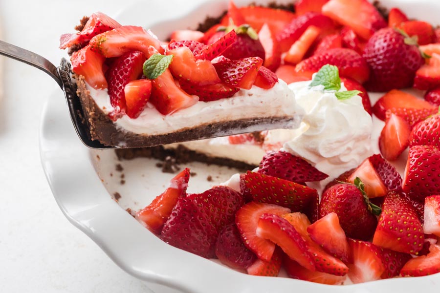 A pie server lifting a slice of strawberry cream pie from a full pie filled with whipped topping and fresh strawberries.