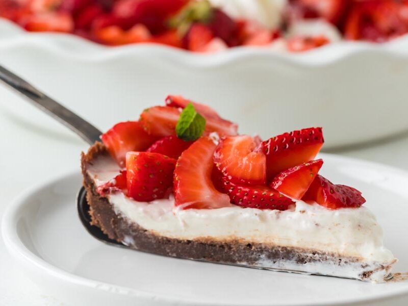 A close-up of a pie slice being served, showing creamy filling, chocolate crust, and sliced strawberries.