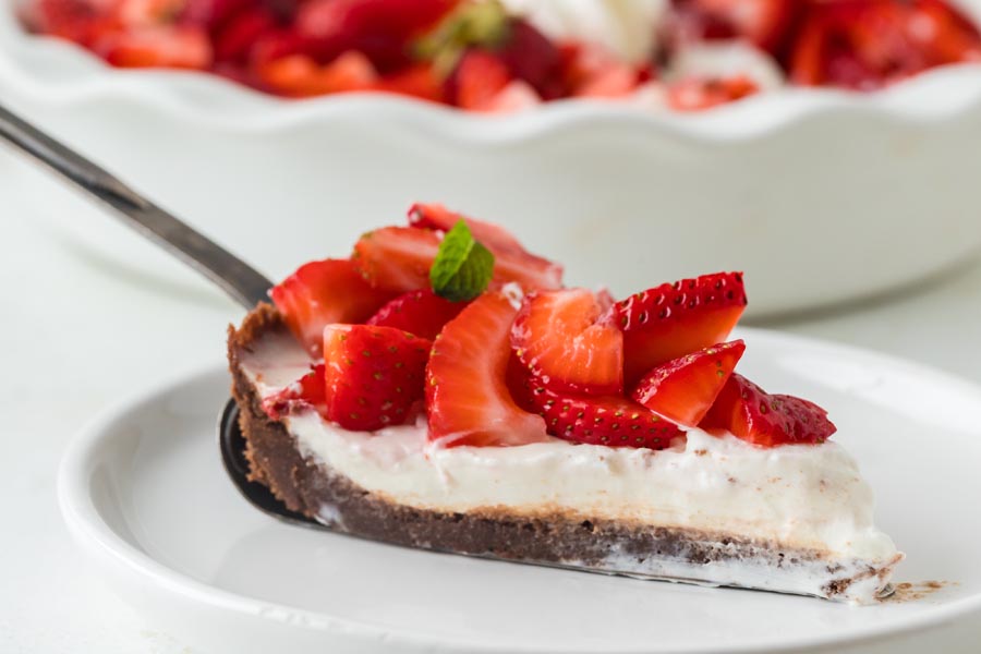 A close-up of a pie slice being served, showing creamy filling, chocolate crust, and sliced strawberries.