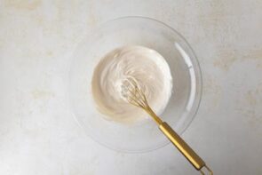Whipped sour cream being prepared in a glass mixing bowl with a gold whisk.