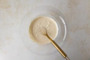 Cream mixture whisked in a glass bowl with a gold whisk, mid-process.