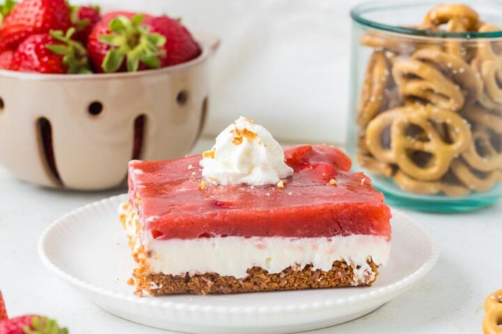 A slice of low-carb strawberry pretzel salad topped with whipped cream sits on a white plate with bowls of strawberries and pretzels in the background.