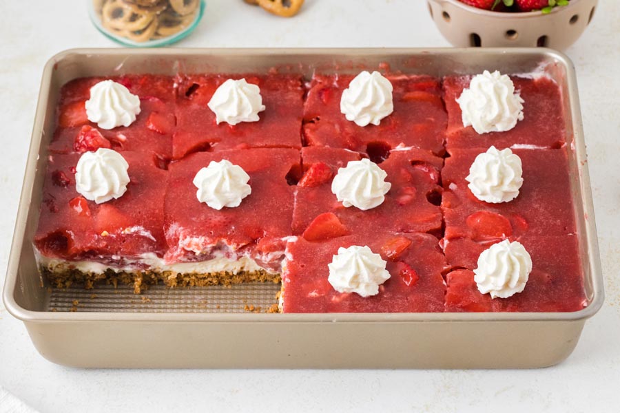 A baking pan of sliced strawberry pretzel salad topped with dollops of whipped cream, with one corner piece missing.