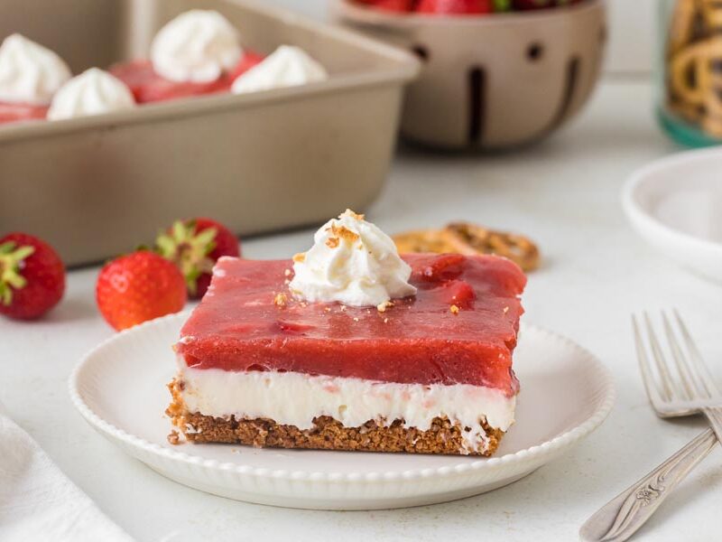 A close-up of a single slice of strawberry pretzel salad on a plate, with the full pan of dessert blurred in the background.