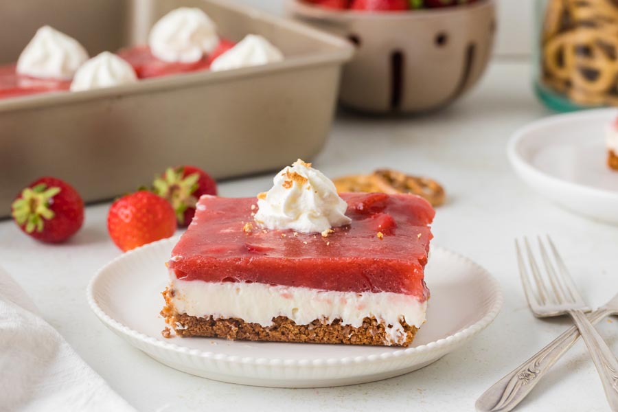 A close-up of a single slice of strawberry pretzel salad on a plate, with the full pan of dessert blurred in the background.