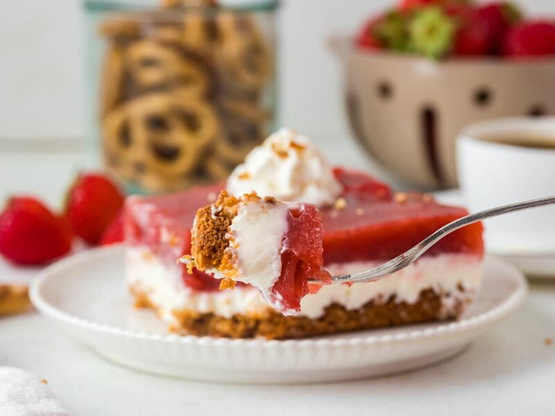 A forkful of strawberry pretzel salad hovers over a plated slice, showing layers of strawberry, cream cheese, and pretzel crust.