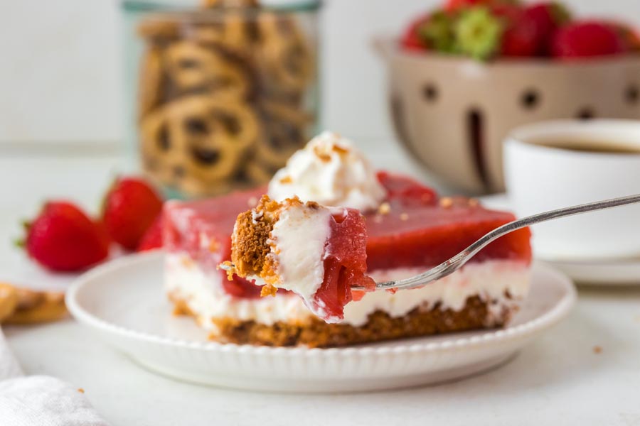 A forkful of strawberry pretzel salad hovers over a plated slice, showing layers of strawberry, cream cheese, and pretzel crust.