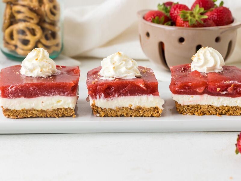 Three slices of keto strawberry pretzel salad topped with whipped cream are arranged in a row on a white serving platter.