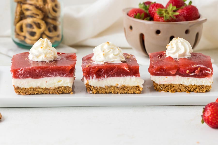 Three slices of keto strawberry pretzel salad topped with whipped cream are arranged in a row on a white serving platter.