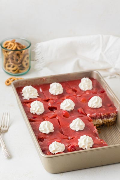 A pan of strawberry pretzel salad cut into squares with whipped cream on top, set beside a jar of pretzels and a white napkin.