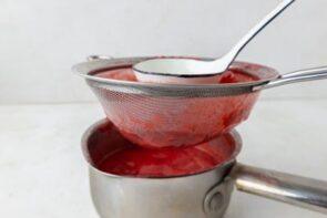 Cooked strawberries being strained through a mesh sieve into a saucepan.