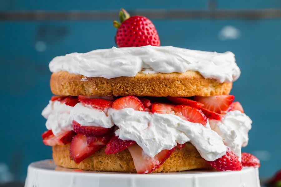 a whole keto strawberry shortcake sitting on a white cakestand with blue background with a strawberry on top