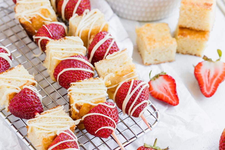 chunks of cake and fresh strawberries on a wood skewer and topped with white frosting on a rack with sliced strawberries near by