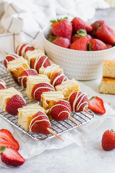 three dessert kebabs with cake and strawberries on a wire rack next to a bowl of strawberries and chunks of cake