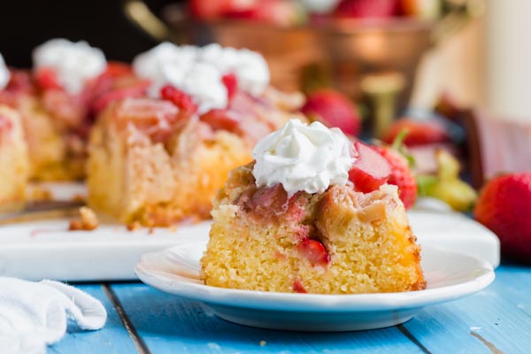 a slice of keto strawberry rhubarb cake on a white plate with a whole cake in the background and strawberries