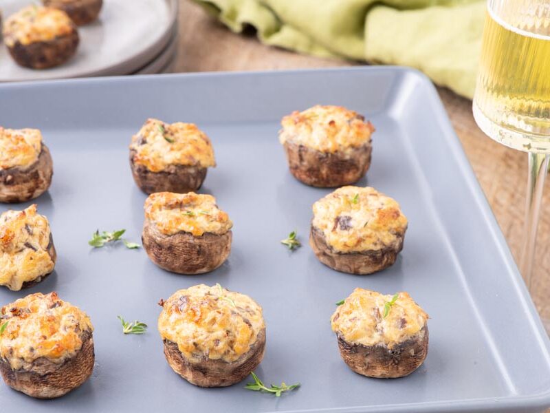 Rows of baked stuffed mushrooms on a baking tray next to a glass of white wine.
