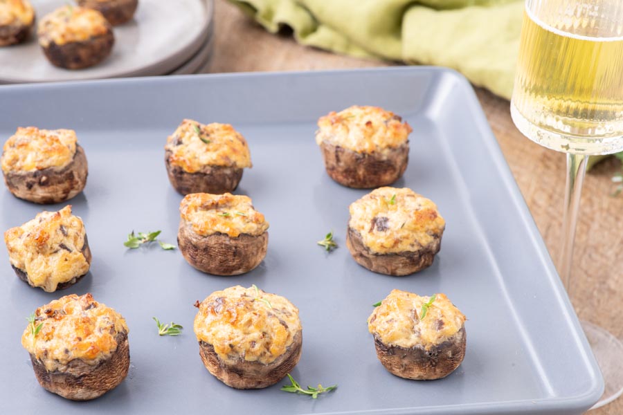 Rows of baked stuffed mushrooms on a baking tray next to a glass of white wine.