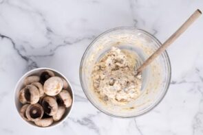A wooden spoon stirring a creamy cheese mixture of sausage next to a bowl of mushroom caps.