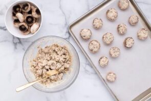 Unbaked stuffed mushrooms on a tray next to a bowl of filling and a bowl of mushroom caps.