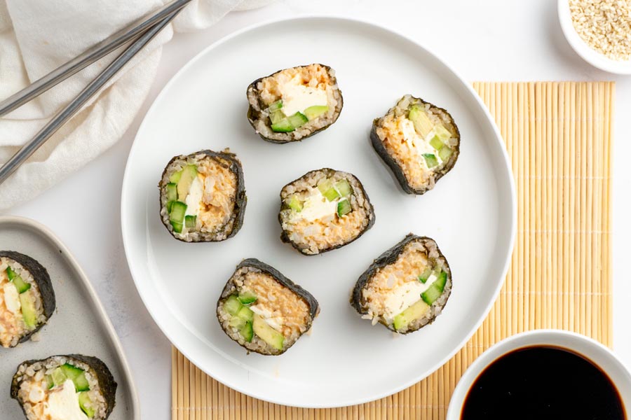 Keto California rolls on a white plate, filled with cream cheese, crab and vegetables. Plate is over a bamboo mat and a bowl of soy sauce is nearby.