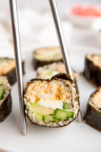 Metal chopsticks holding a sushi roll upright to show the layers of crab, cream cheese, avocado and cucumber.