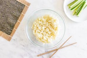 Cauliflower rice in a bowl next to chopsticks, nori and sliced cucumber.