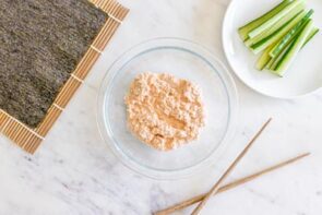 Crab meat in a clear bowl next to chopstick, nori sheet over bamboo mat and sliced cucumber.