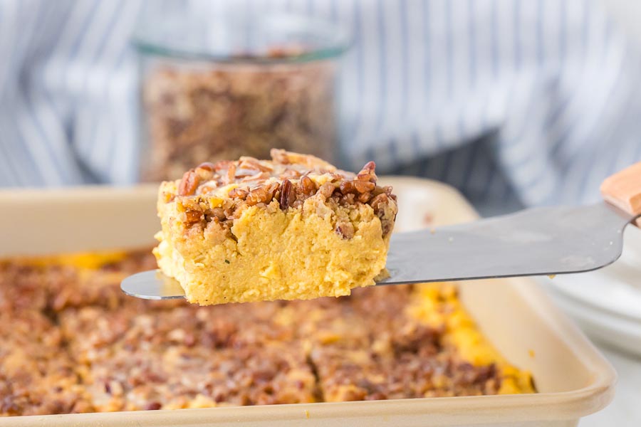 A close-up of a single square slice of keto sweet potato casserole being lifted from the baking dish, showing its fluffy pumpkin-cauliflower interior and pecan topping.