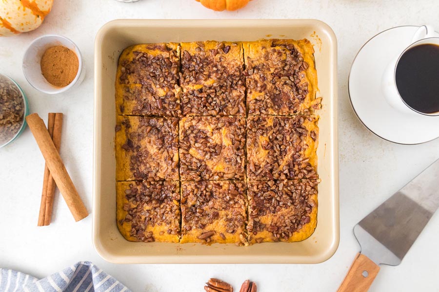 Overhead view of a full pan of keto sweet potato casserole sliced into nine servings, surrounded by cinnamon sticks, a cup of coffee, and fall decor.
