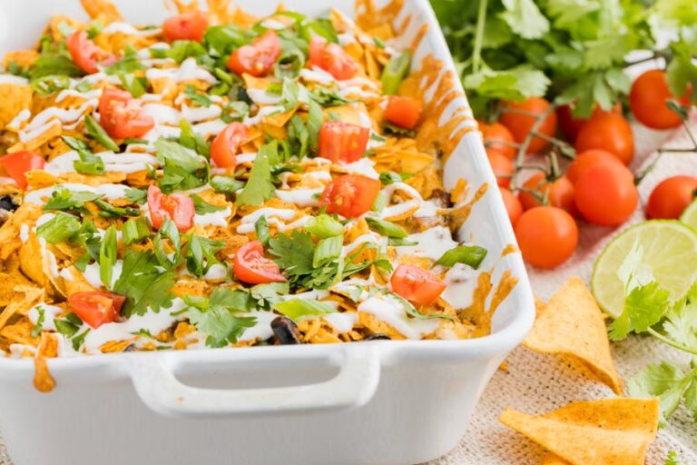 taco casserole in a white baking dish topped with crushed tortilla chips, cilantro and tomatoes next to tortilla chips and limes