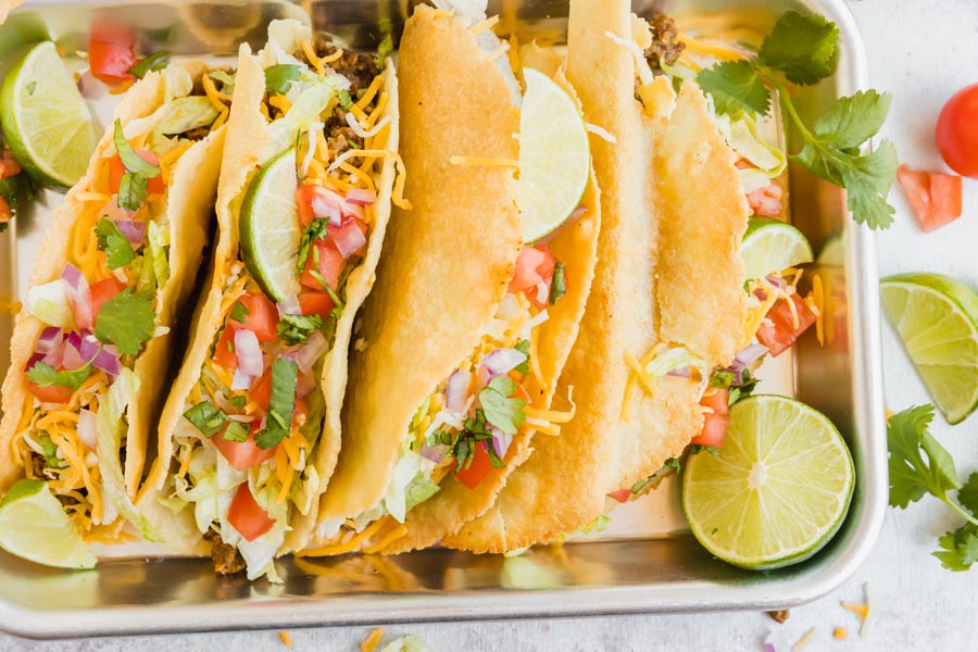 four hard shell tacos with beef, cheese, cilantro and a slice of lime near by