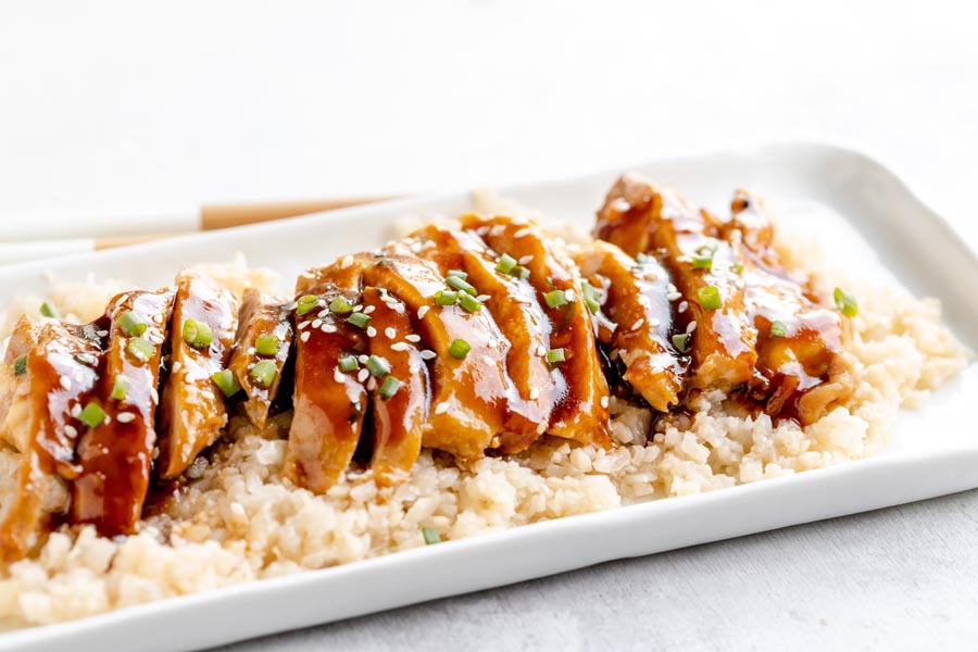 sliced pieces of teriyaki chicken over a bed of cauliflower rice and topped with green onion and sesame seeds