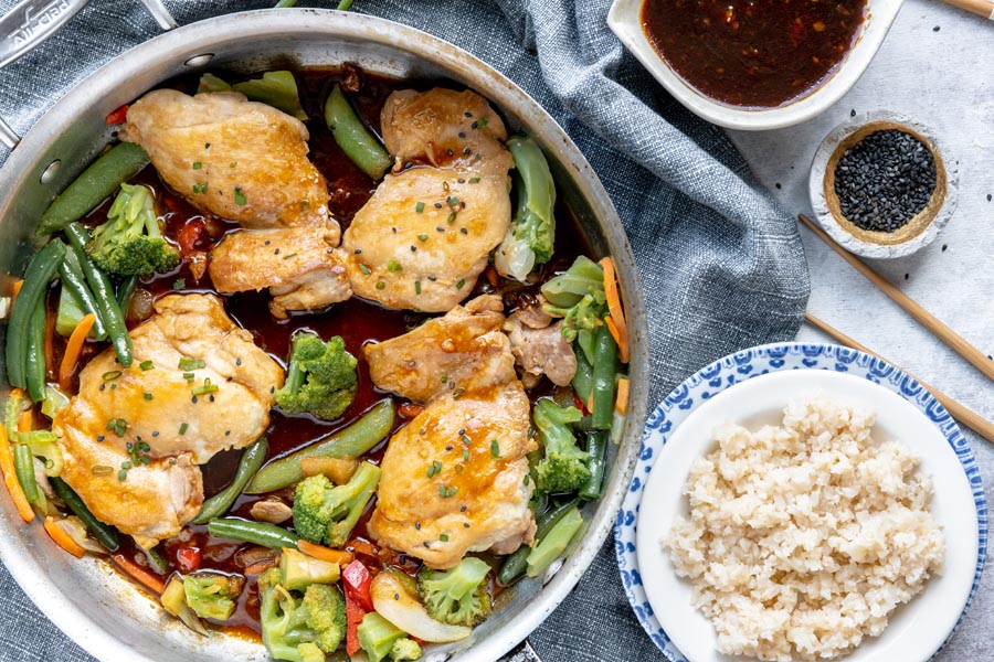 a skillet with cooked teriyaki chicken with mixed stir fry vegetables around next to a plate with cauliflower rice and chopsticks