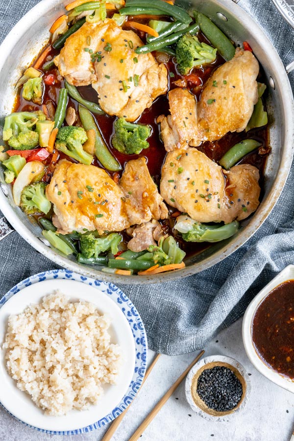 a skillet with four pieces of chicken thighs coated with sauce and topped with black sesame seeds sits next to a plate with cauliflower rice and more black sesame seeds