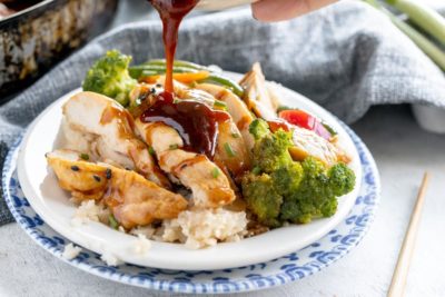 pouring teriyaki sauce on sliced chicken and stir fry vegetables on a white plate. a chopstick sits nearby