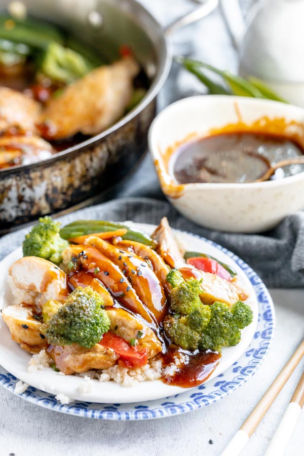 a plate with teriyaki chicken and vegetables with a bowl of teriyaki sauce in the background and a skillet with chicken