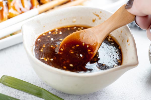 holding a small wooden spoon in a bowl of amber colored teriyaki sauce with sesame seeds