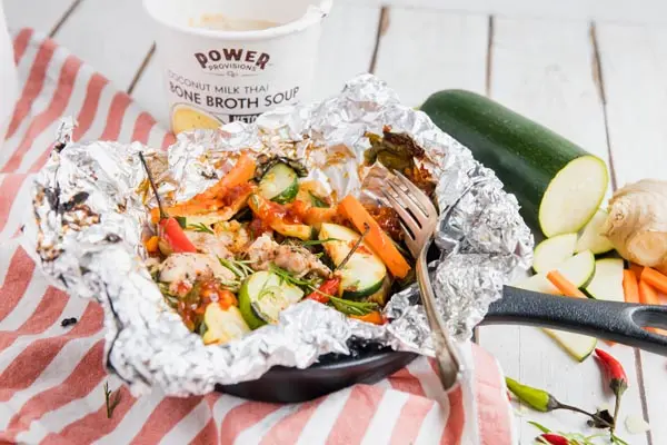 a chicken camping foil packet in a castiron skillet with vegetables and a cup of soup nearby