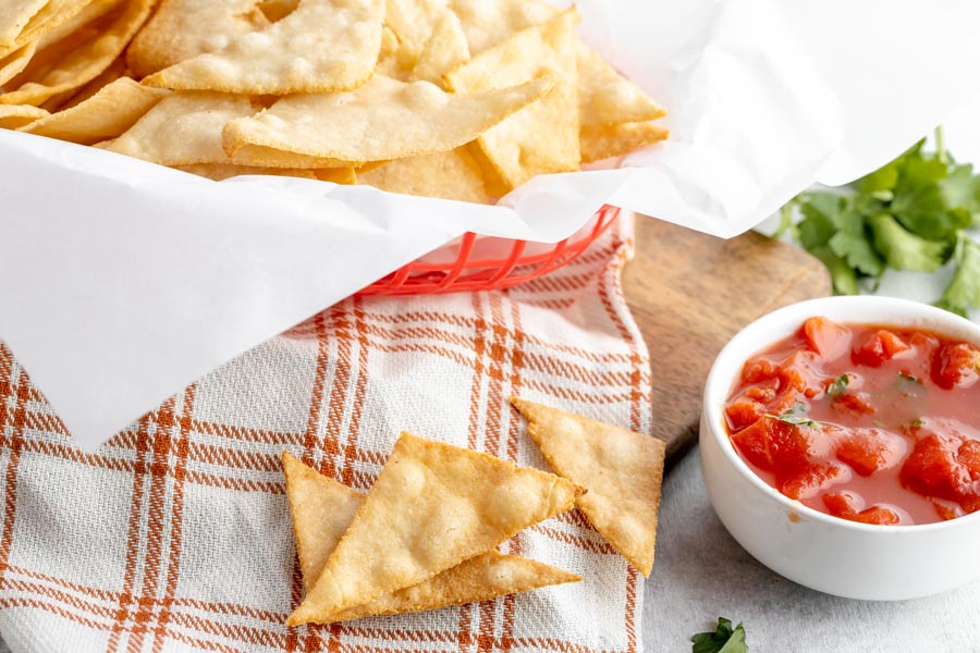 three tortilla chips sit on a checkers towel next to red salsa with a basket of chips behind